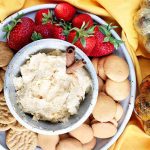 Top-down shot of a chip and dip set filled with pumpkin cream cheese garnished with two cinnamon sticks, surrounded by cookies and strawberries, on a yellow cloth with one silver and one gold decorative pumpkin.