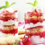Two glass jars of strawberry yogurt parfait, garnished with green mint, on a red and white checkered tablecloth with scattered whole berries and lemon cornmeal cookies, on an off-white background.