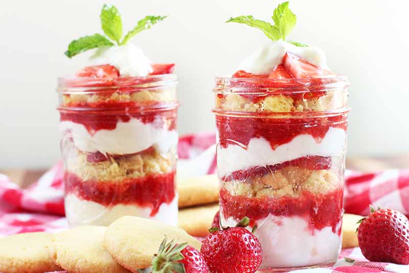 Two glass jars of strawberry yogurt parfait, garnished with green mint, on a red and white checkered tablecloth with scattered whole berries and lemon cornmeal cookies, on an off-white background.