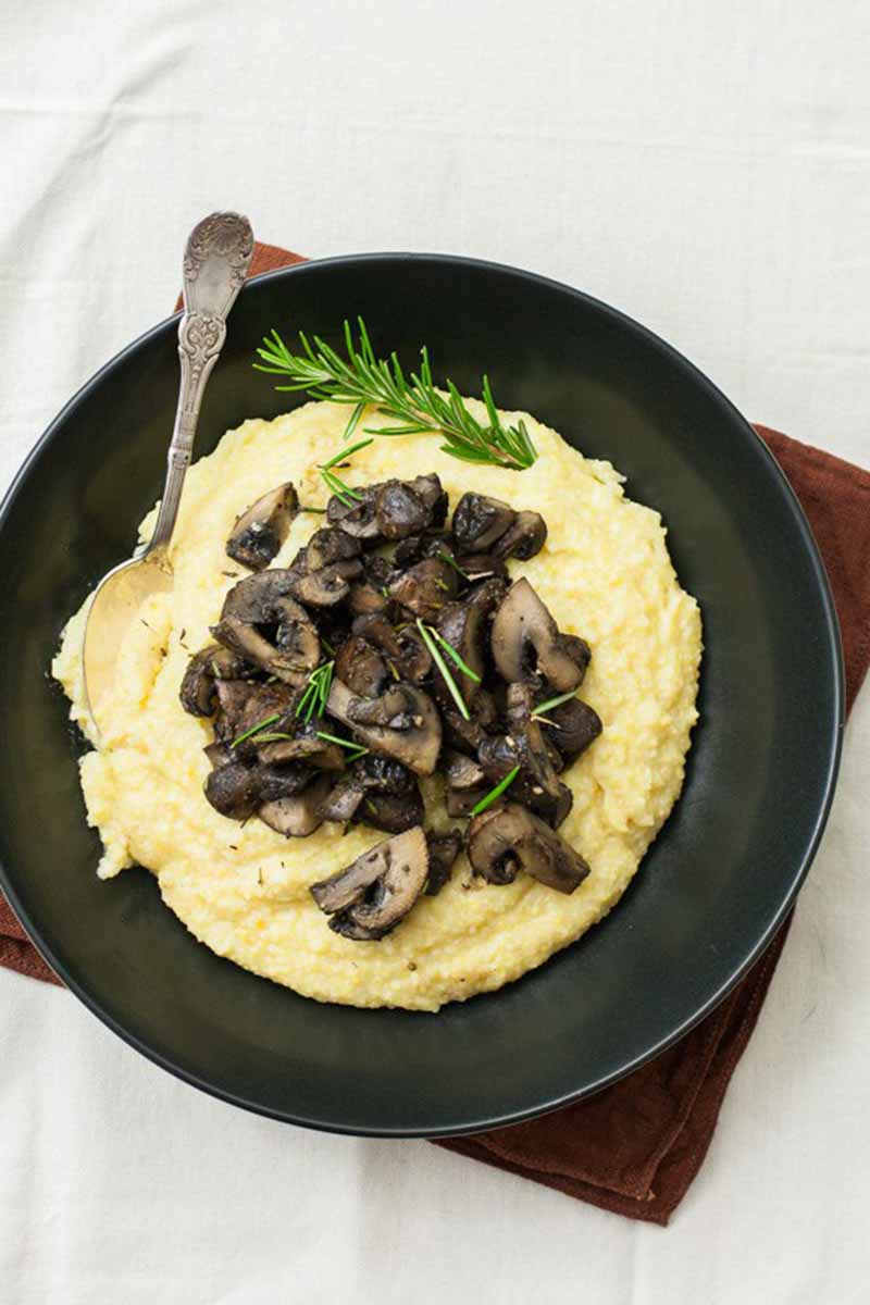 Vertical image of a bowl with creamy polenta and baby bellas with rosemary and a spoon.