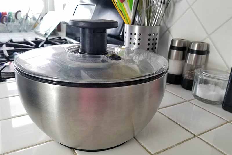Une essoreuse à salade OXO en acier inoxydable et en plastique noir sur un comptoir de cuisine en carreaux blancs, avec des moulins à sel et à poivre, une boîte à ustensiles, une cave à sel en verre et une cuisinière à gaz.