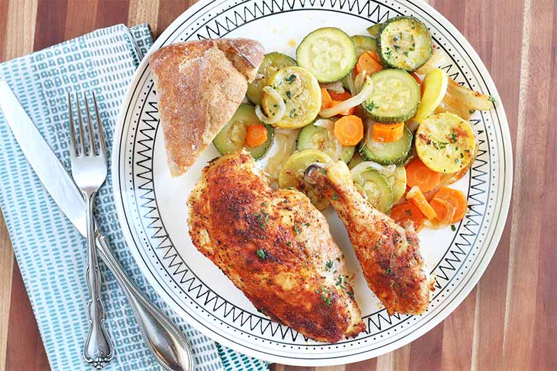 A roasted spiced chicken breast and drumstick on a black and white patterned plate with a triangle of crusty bread, squash, zucchini, and carrots, on a striped brown table with a folded blue and white napkin and silverware.