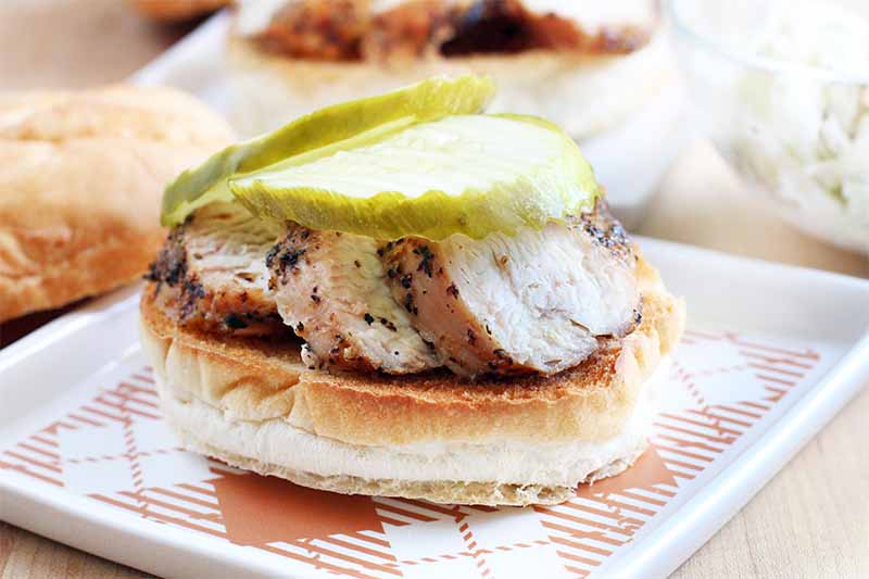 Sliced grilled chicken coated with spices and topped with two large pickle slices is on a square white and orange plate, with the top of the roll and another sandwich in the background, on a beige particleboard surface.