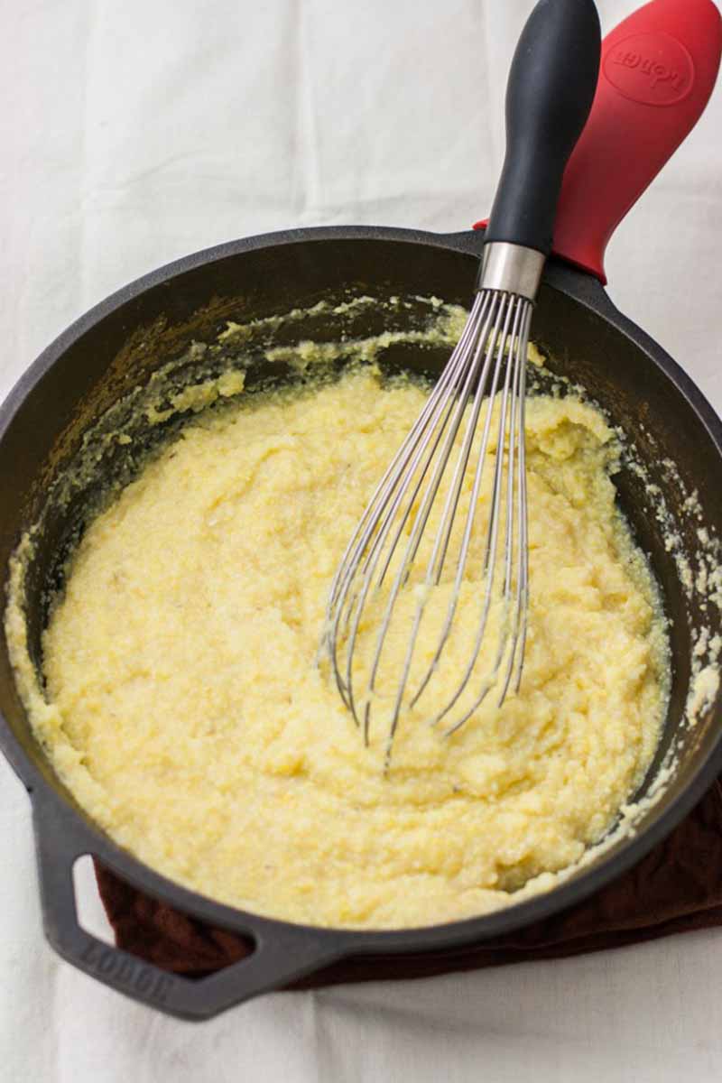 Vertical image of a pot with cooked cornmeal and a whisk on a white cloth.