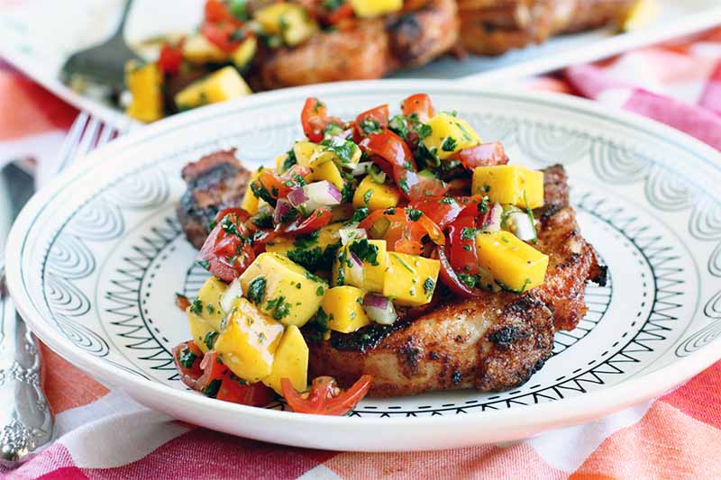 Caramelized grilled bone-in porkchop on a white dinner plate with a black geometric design, topped with mango, cilantro, and tomato salsa, on a red, orange, and white checkered tablecloth with silverware and another plate in the background.