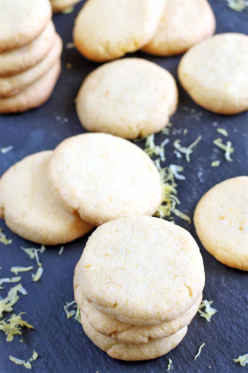 Round beige cookies on a black slate background with scattered lemon zest.