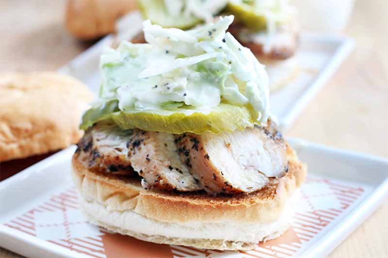 Half of a kaiser roll is topped with sliced chicken, pickles, and slaw, on a square white and orange plate, with the other half of the bun and another sandwich in the background.