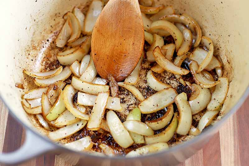A wooden spoon stirs sliced onions in an enameled pot.