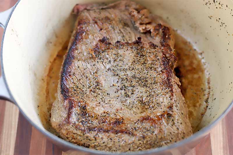 Browned and seasoned brisket in a large enameled pot, on a wood surface.