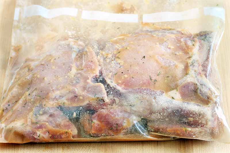 Pork chops marinating in a garlic and herb mixture in a large zip-top plastic bag, on a beige kitchen counter.