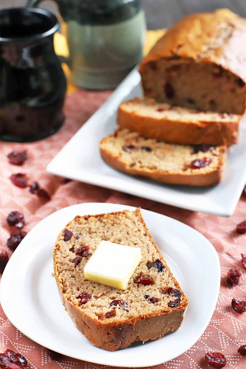 A slice of homemade cranberry bread topped with a pat of butter on a white plate, with more in the background on a rectangular white serving platter, on a pink tablecloth with scattered dried berries, beside green and black ceramic mugs.