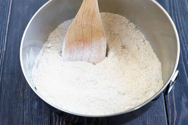 A wooden spoon stirs flour and other dry ingredients in a stainless steel bowl.