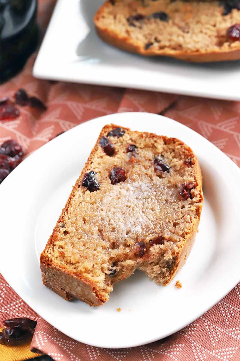 A slice of cranberry bread topped with melted butter with a bite taken out of it, on a white plate, with a rectangular serving platter of more in the background, on a pink patterned tablecloth topped with scattered dried berries.