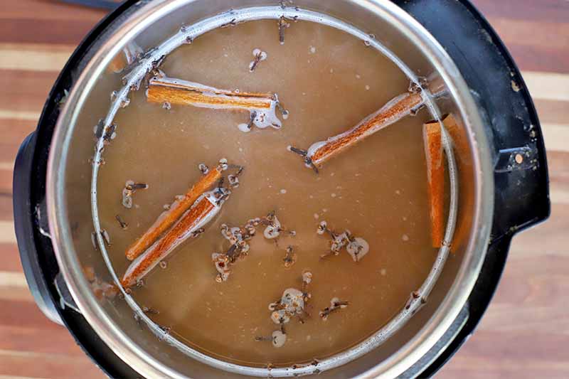 Overhead shot of a slow cooker full of apple cider with whole cinnamon sticks and cloves, on a wooden surface.