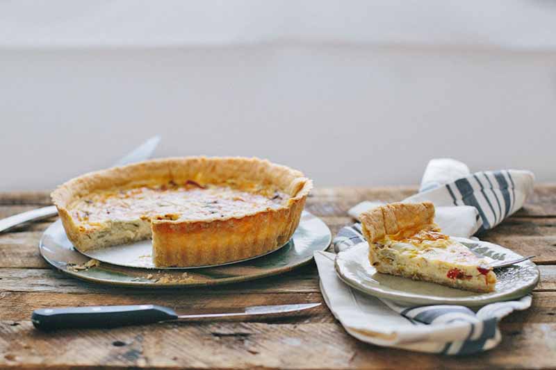 Horizontal image of a quiche with a golden crust, and a slice removed on a plate, all on a wooden table.