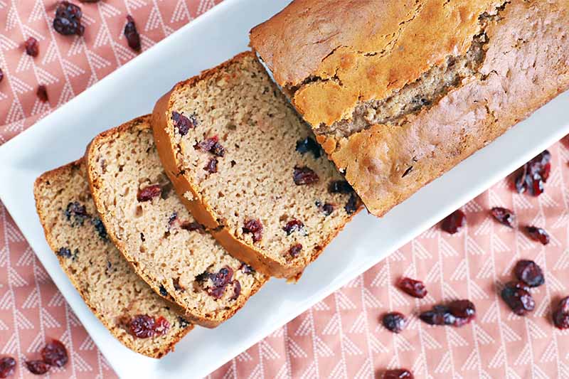 Overhead shot of a loaf of cranberry quick bread and three slices arranged on a rectangular white ceramic serving platter, on a patterned pink tablecloth topped with scattered dried berries.