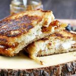 A golden brown grilled cheese sandwich, cut in half and stacked on a wooden log slice serving board, on a wood background with a jar of fig jam and dried fruit.