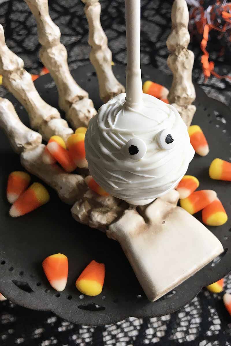Vertical image of a mini mummy dessert ball on a stick on a skeleton hand with candy corn decorations.