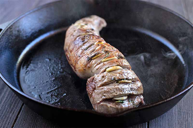A pork tenderloin with garlic and herbs inserted in slits running down the length of the meat is browning in a large oiled cast iron pan.