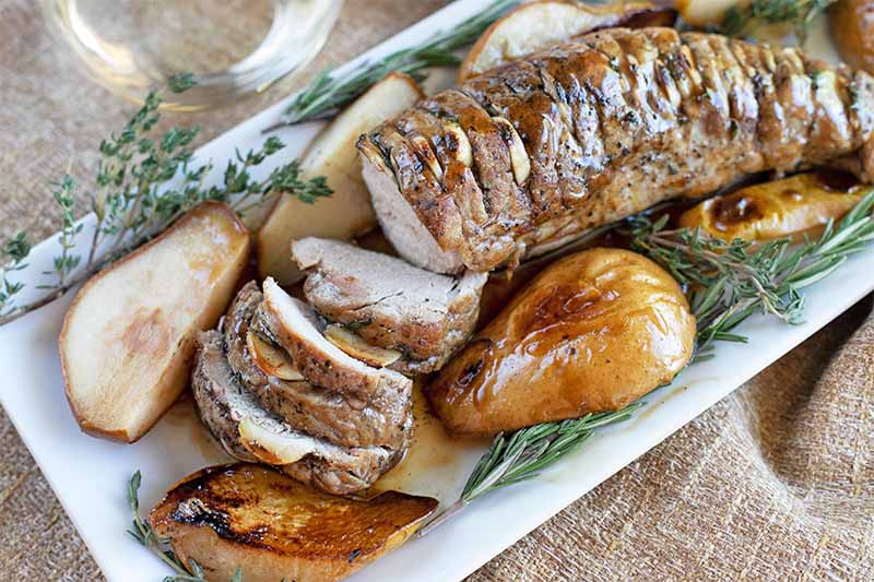 Partially sliced roasted pork tenderloin with pears, rosemary, and thyme, on a rectangular white ceramic serving platter, on a burlap surface.
