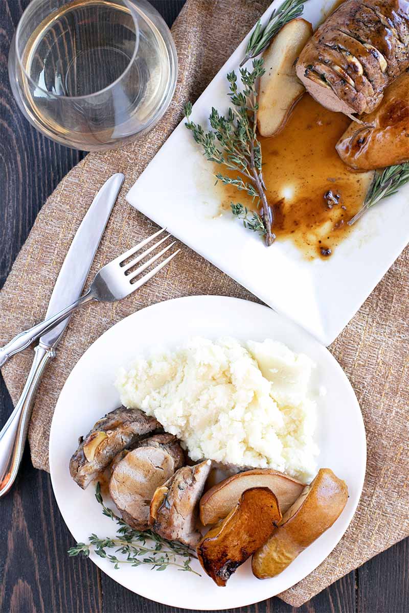 Overhead shot of a round white plate and a rectangular serving dish of pork tenderloin with pears, mashed potatoes, fresh herbs, and brown gravy, on brown burlap with a fork and knife, and a glass of white wine.