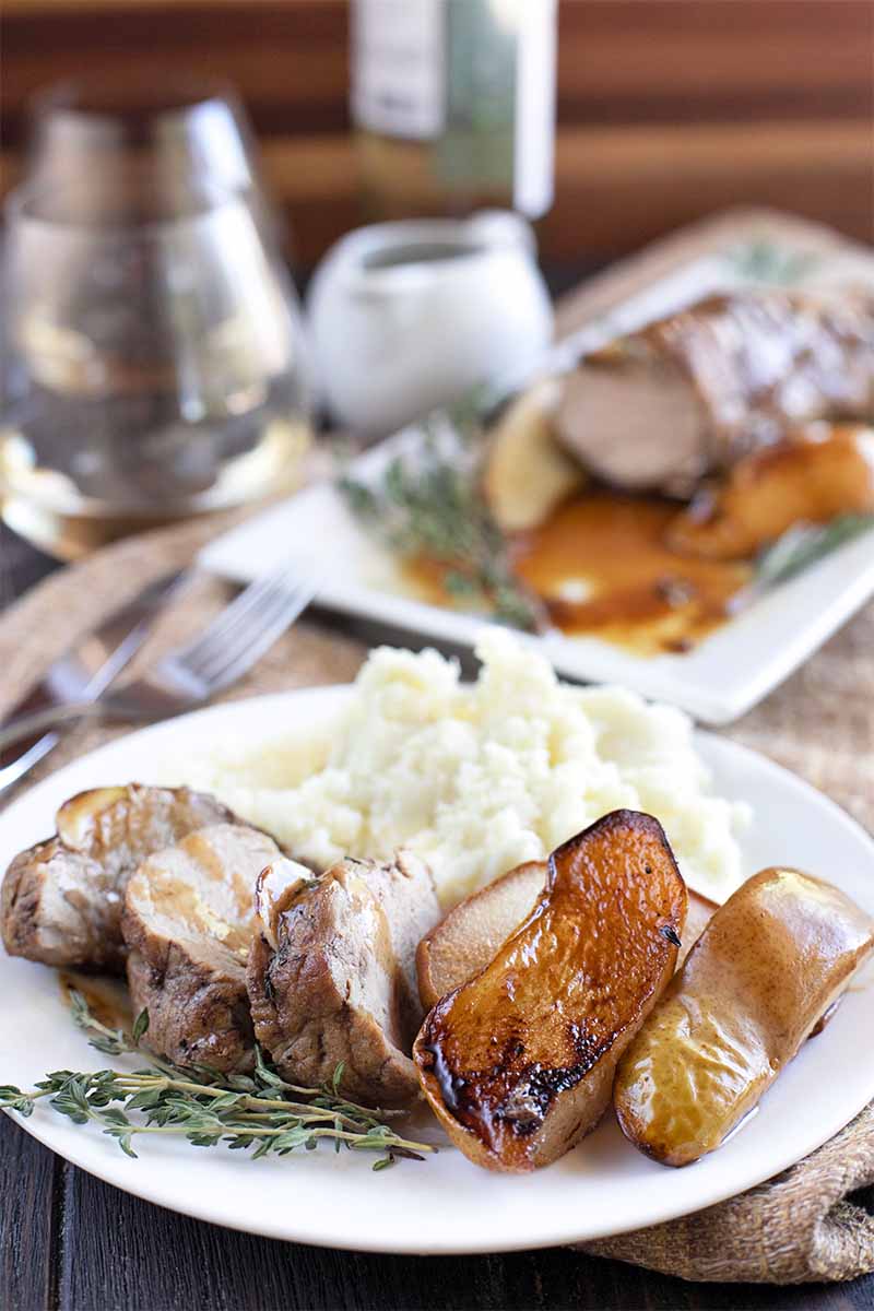 Vertical shot of a white ceramic plate of sliced pork tenderloin, pears, and mashed potatoes, with a rectangular serving platter of more of the meat, wine glasses, and utensils in shallow focus in the background.
