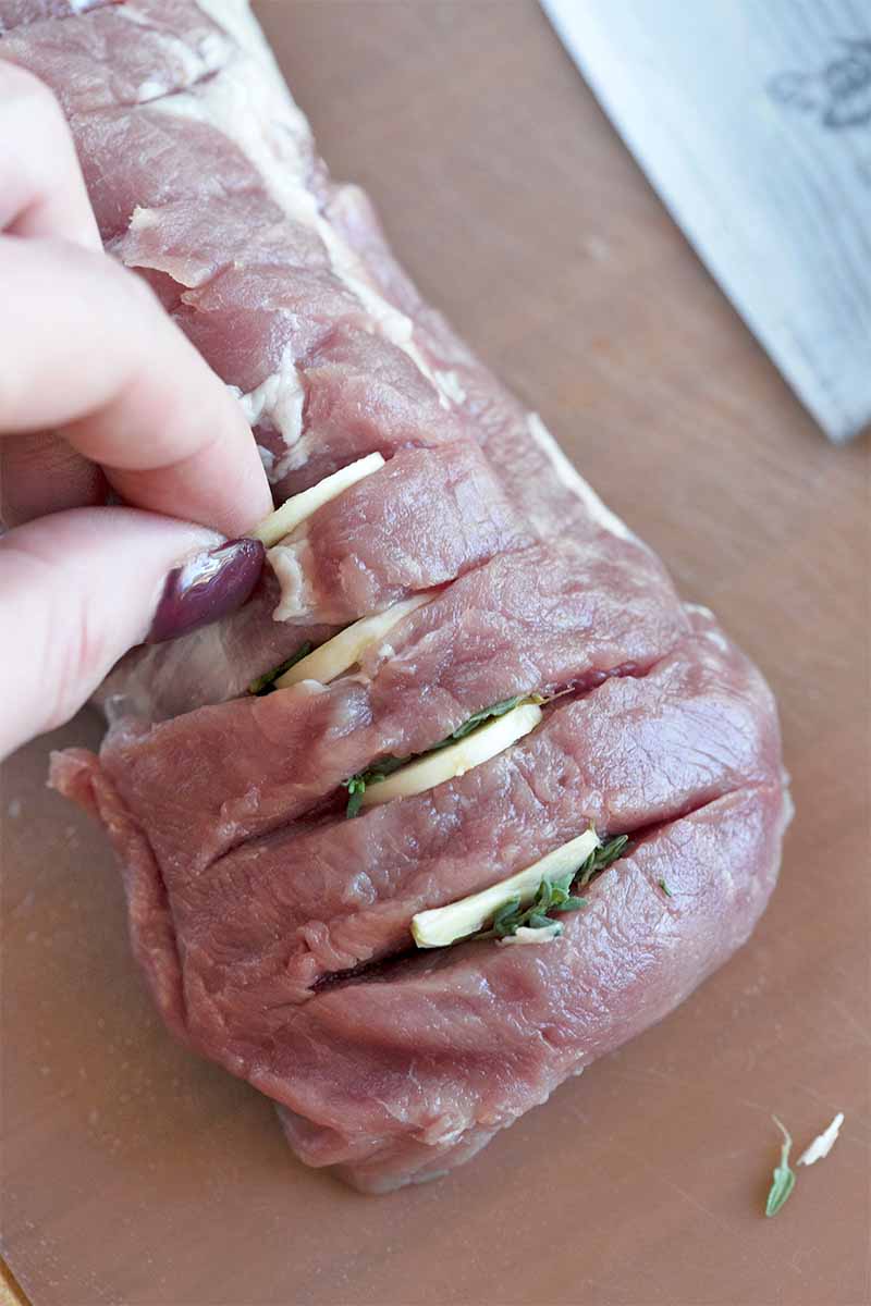 A hand inserts slices of garlic and fresh herbs into slits cut into an uncooked pork tenderloin.