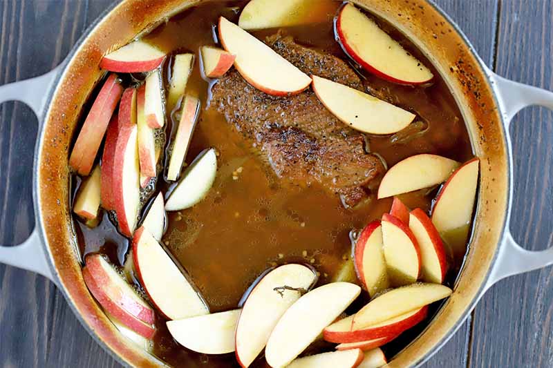 Top-down shot of sliced apples with brisket and a brown liquid in a large cooking pot, on a dark brown wood surface.