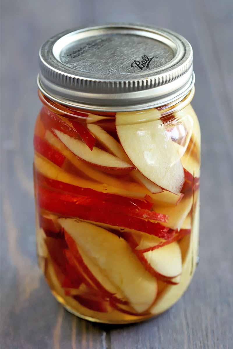 A sealed mason jar of sliced apples in brandy, on a wood background.