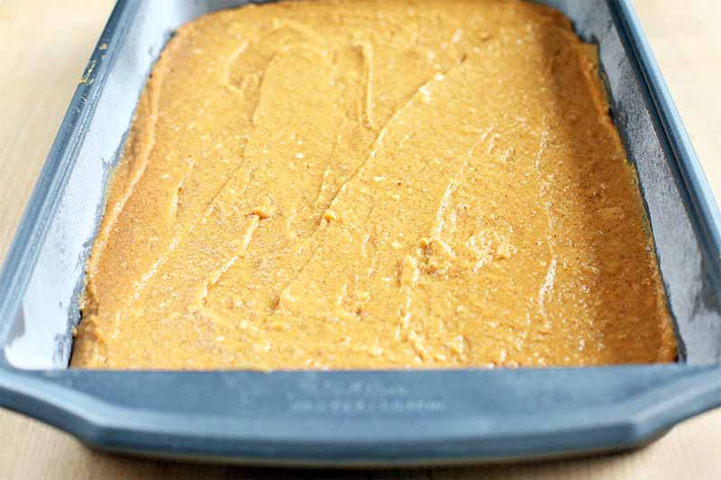 Orange-colored cake batter is spread smooth on top, filling a large square metal baking pan coated with oil and flour, on a beige background.