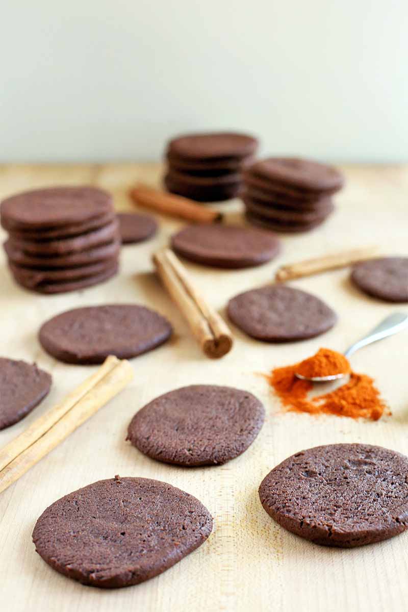 Vertical image of chocolate cookies, cinnamon sticks, and a scattered spoonful of cayenne pepper, on a beige surface against an off-white wall.