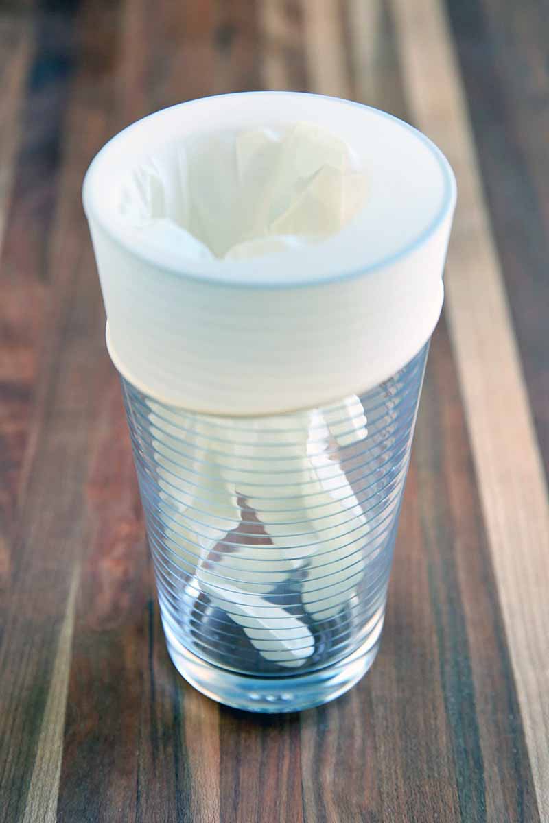 A white disposable glove is suspended in a tall drinking glass, with the wristband wrapped over the rim of the glass, on a brown striped surface.