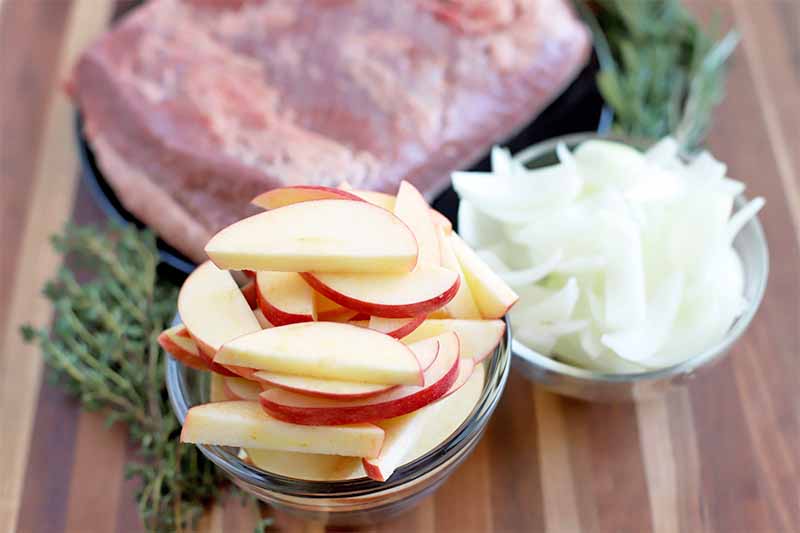 Small glass bowls of sliced apples and onions in front of a black plate of raw brisket and sprigs of fresh herbs, on a brown wood surface.