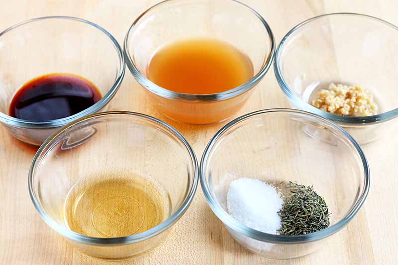 Five small glass bowls of soy sauce, apple cider vinegar, minced garlic, dried oregano, salt, and vegetable oil, on a beige wood countertop.