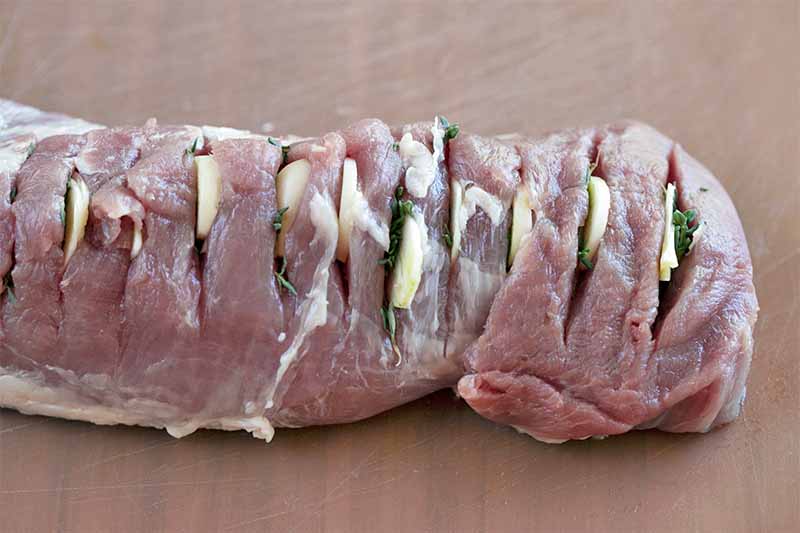 Raw pork tenderloin with slices of garlic and fresh herbs inserted in slits across the length of the meat, on a beige wood surface.