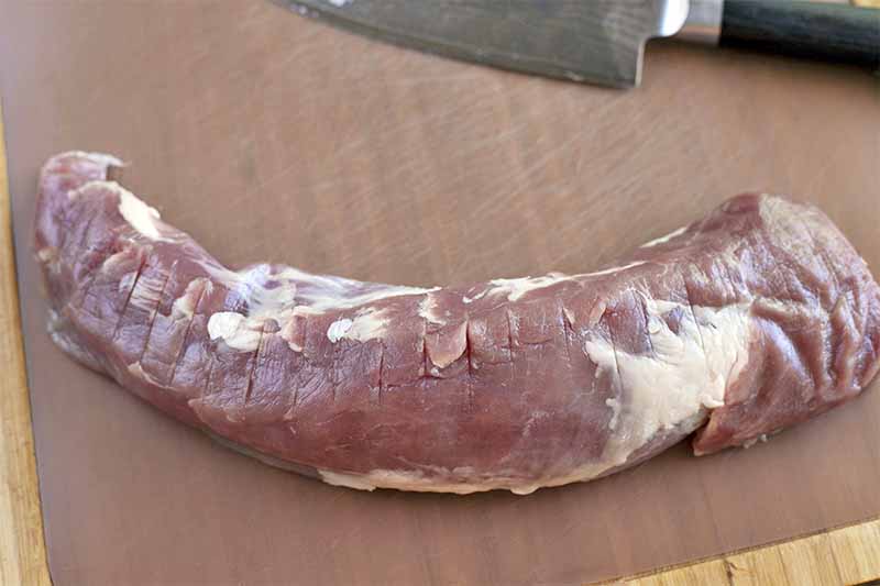 A pork tenderloin with slits cut into it across the full length of the meat, with a chef's knife on a wood surface.