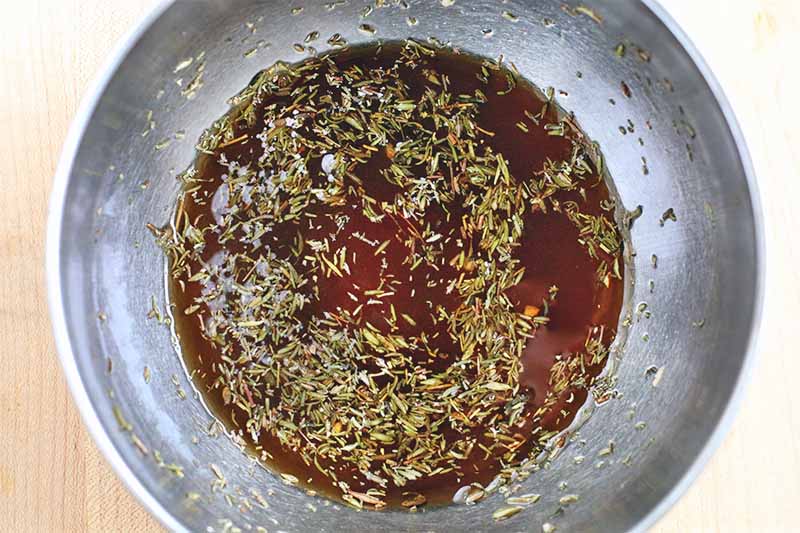 Top-down shot of a marinade mixture for pork, topped with dried thyme, at the bottom of a large stainless steel bowl on a beige counter.