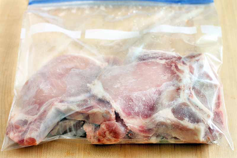Bone-in pork chops marinating in a zip-top plastic bag on a beige countertop.