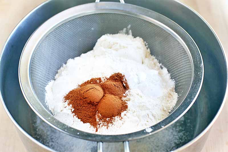Cinnamon and flour are being sifted through a fine mesh strainer set into a stainless steel mixing bowl, on a beige background.