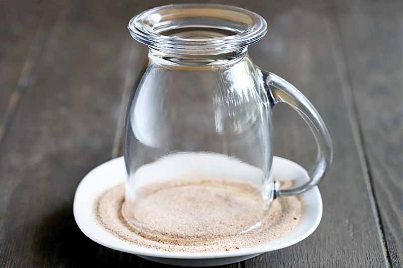 A glass mug with a handle is inverted onto a saucer filled with cinnamon sugar, on a dark brown wood surface.