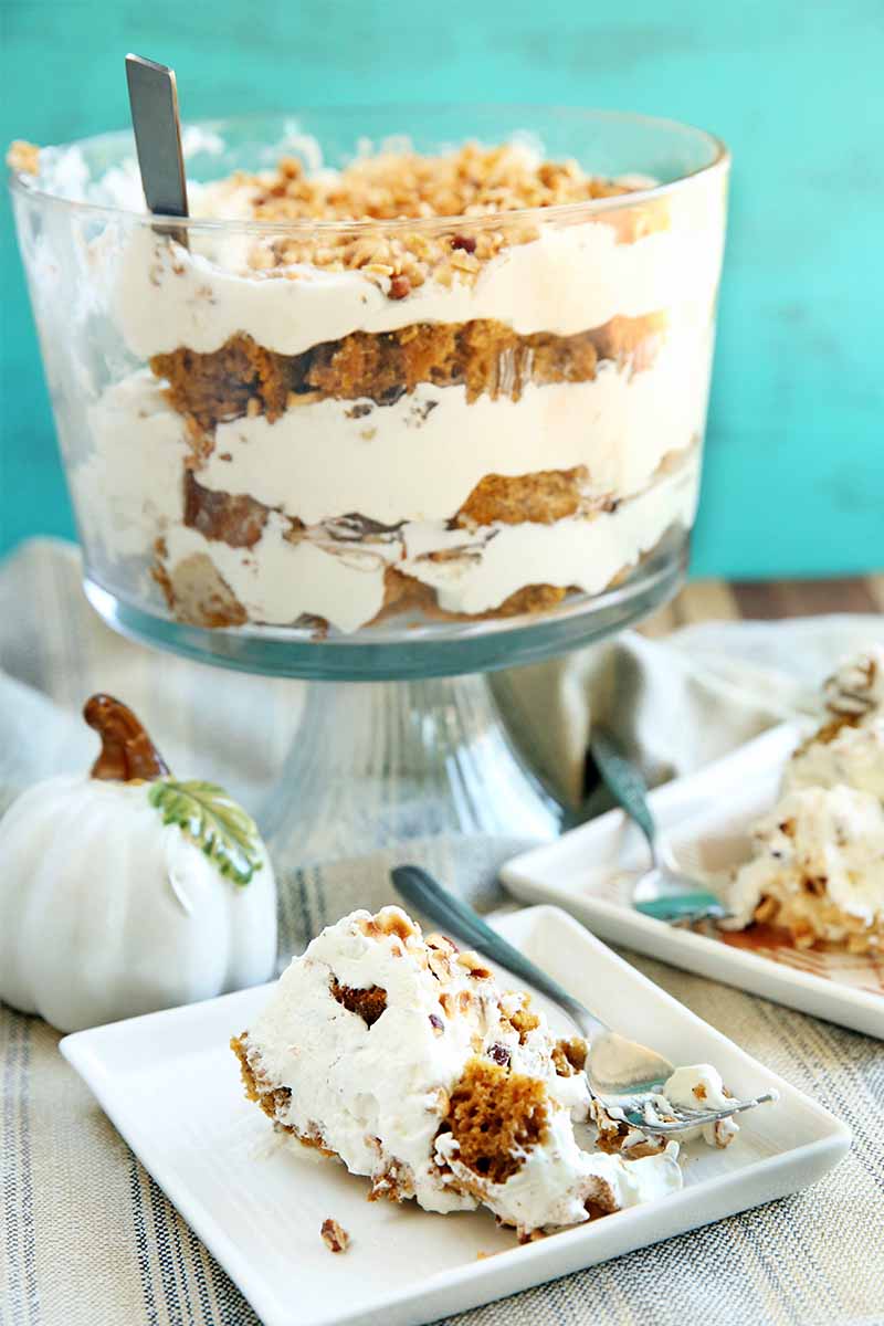 Vertical image of layered pumpkin trifle in a large glass serving dish with pedestal, with a white ceramic pumpkin and two white square plates of the dessert with forks, on a striped blue and gray cloth, with a mottled aqua background.