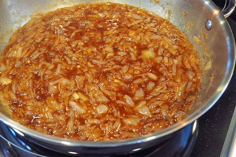Onions cooking in a red sauce in a large stainless steel frying pan on a stove.