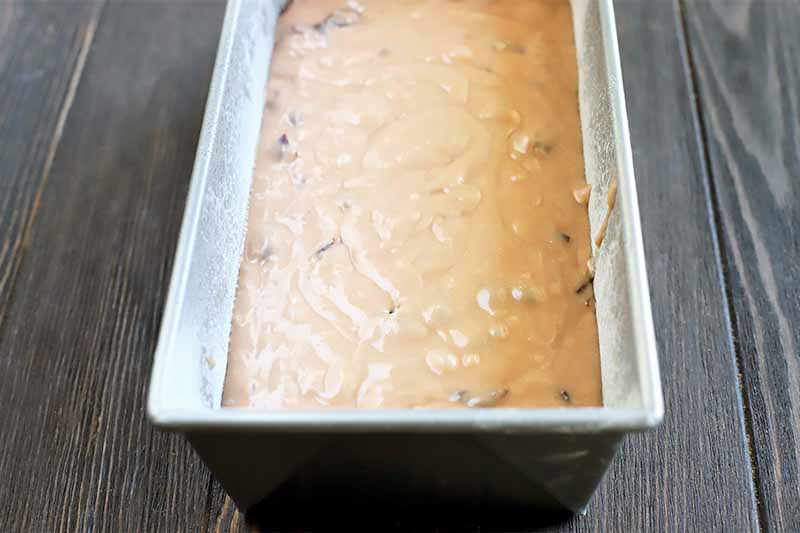 A beige quick bread batter dotted with dried cranberries fills a metal loaf pan, on a brown wood surface.