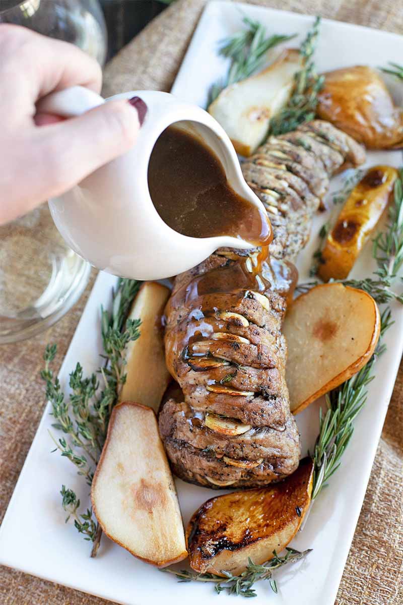 A hand pours sauce from a small white pitcher onto a Hasselback-style garlic and herb pork tenderloin with roasted pears, on a square white ceramic serving dish, on a burlap fabric surface.