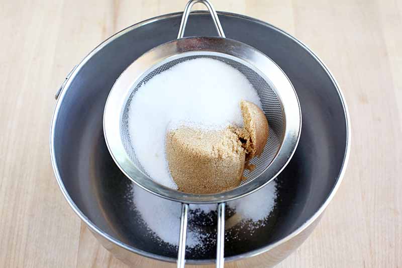 A small metal mesh strainer is being used to sift white and packed brown sugar into a stainless steel bowl, on a light beige wooden countertop.