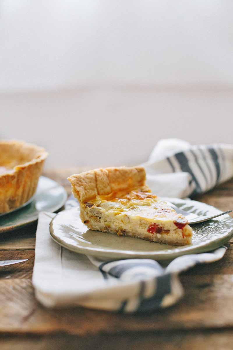 Vertical image of a slice of quiche on a plate with a fork on it, over a striped white towel.
