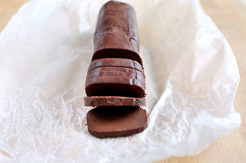 A log of chocolate cookie dough is sliced into rounds, on a crumpled piece of waxed paper, on a beige background.