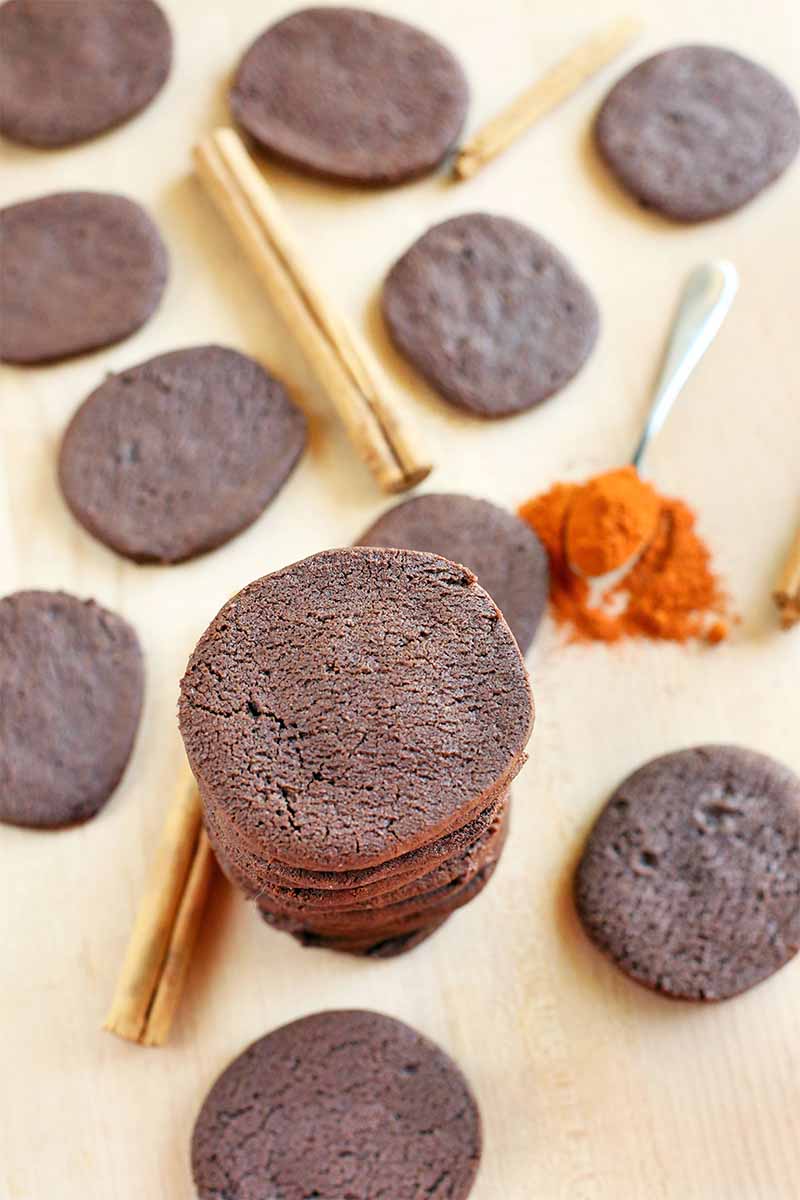 Top-down vertical shot of a tall stack and scattered chocolate cookies, with whole brown cinnamon sticks and an overfilled spoonful of orange cayenne pepper powder, on a beige surface.