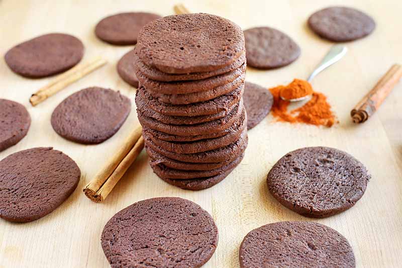 Chocolate cookies are scattered on a beige wooden surface, with a spoonful of cayenne pepper, cinnamon sticks, and a tall stack of cookies in the middle.
