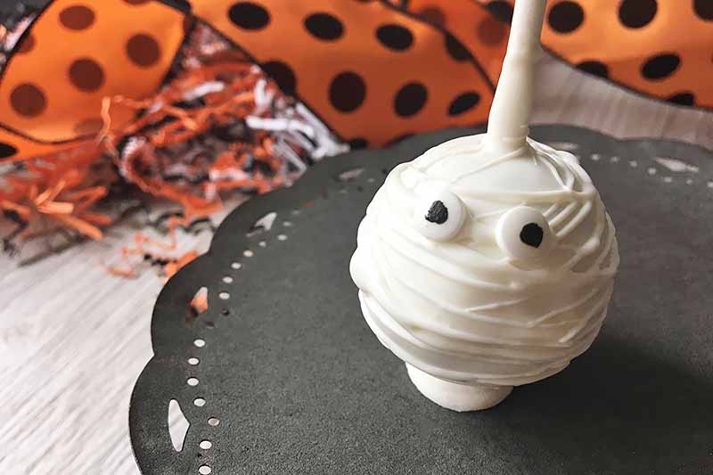 Horizontal image of one mummy cake pop on a black plate in front of orange and black ribbons.